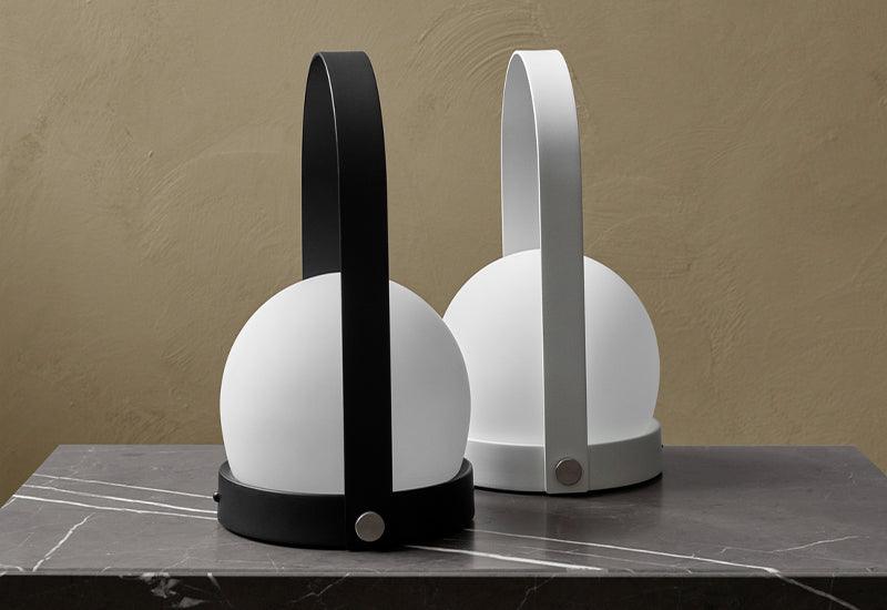 Two Carrie Portable portable lamps stood side by side. One is in white and one is in black. Both are designed by NORM architects for Audo Copenhagen.