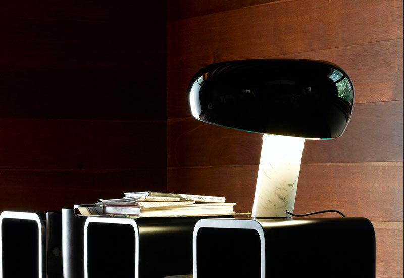  The Snoopy table lamp by A Castiglioni & P.G Castiglioni for Flos qith a black glass shade and a white marble base, placed on a shelving unit with magazines.